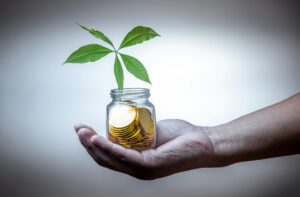 Hand met glazen pot vol munten waar een groene plant uit groeit, symbool voor duurzaam investeren en financiële groei